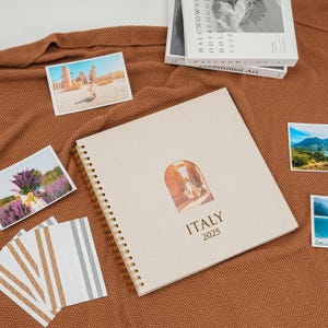 Può includere: Un diario a spirale color crema con la scritta "ITALY 2025" sulla copertina, con una finestra ritagliata che mostra una fotografia. Diverse piccole fotografie e una pila di carte sono disposte attorno al diario su una superficie marrone.