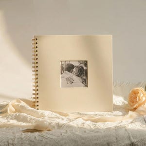 May include: Cream-colored photo album with a square cutout displaying a black and white photo of a couple embracing. The album has a gold spiral binding. A peach-colored rose and cream-colored fabric are in the foreground.