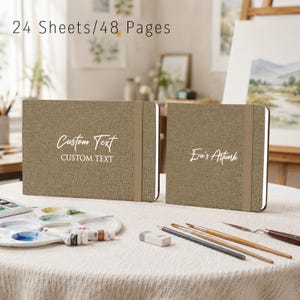 May include: Two rectangular sketchbooks with tan fabric covers and elastic closure bands. The left sketchbook features the white text "Custom Text" in script and block fonts. The right sketchbook features the white script text "Eva's Artwork". 24 Sheets/48 Pages.