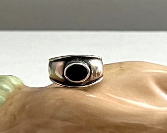 May include: A silver ring with a black stone set in the center. The ring is wide and has a rounded shape.