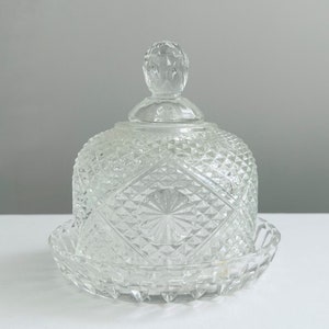May include: A clear glass butter dish with a domed lid and a decorative diamond pattern. The dish has a base that is slightly wider than the lid.