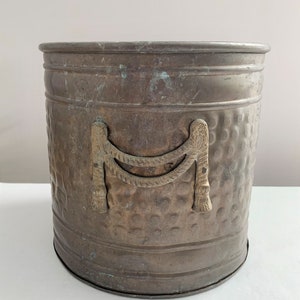 May include: A hammered copper planter with decorative brass handles. The planter has a rounded oval shape and a textured surface.