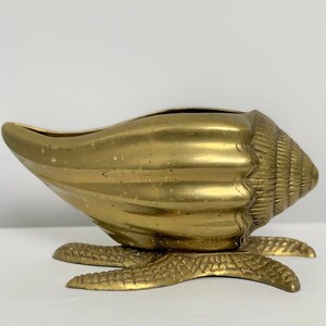 May include: A gold brass seashell planter with a detailed design. The planter is on a base with three legs that resemble a sea creature.