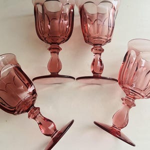 May include: Four vintage pink wine glasses with a decorative design. The glasses have a stem and a wide base. The rims of the glasses have a darker pink trim. The glasses are arranged on a white surface.