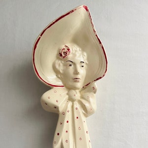 May include: A white ceramic wall plaque featuring a woman's face with a red and white polka dot bow and a large white hat with a red trim. The woman is wearing a red flower on her hair.