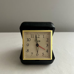 May include: A black and gold travel alarm clock with a white face and black numbers. The clock has a red second hand and a black minute hand. The clock is in a black case.