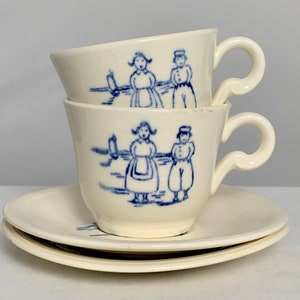 May include: Two cream-colored teacups and saucers stacked, each with a blue illustration of figures in traditional Dutch clothing. The cups have curved handles and the saucers are round. The design is a classic, vintage style.