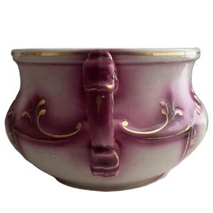 May include: A ceramic bowl with a cream-colored base and deep magenta gradient shading. The bowl features a prominent vertical handle, a gold-painted rim, and delicate gold filigree scrollwork patterns decorating the exterior surface.