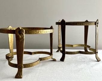 May include: Two gold metal plant stands with ornate designs. The stand on the left has a round top and three legs. The stand on the right has a square top and four legs.