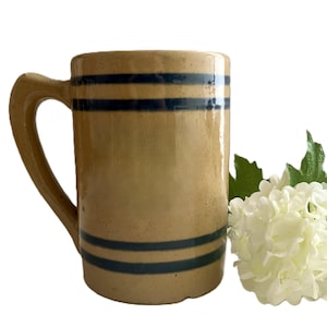 May include: A cylindrical stoneware mug with a tan, glazed finish and a single handle. The mug features two sets of double horizontal dark blue stripes, one set near the top rim and one set near the base, encircling the exterior of the vessel.