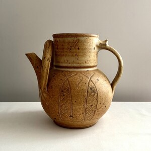May include: A brown ceramic pitcher with a textured surface and a carved design. The pitcher has a handle and a spout.