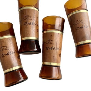 May include: Four amber-colored glass tumblers with a wooden band and gold trim. The wooden band is engraved with the name "Robbin's" and a hat logo. The glasses have a flared base and a rounded top.