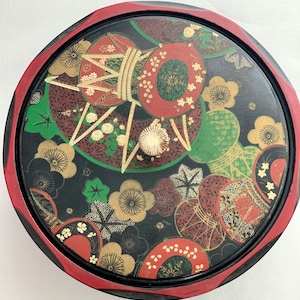 May include: A round, black lacquered box with a red rim and a decorative lid. The lid features a colorful Japanese design with gold accents, including circles, flowers, and geometric patterns.