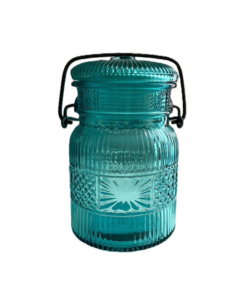 May include: A teal glass jar with a lid and a black metal handle. The jar has a ribbed design with diamond-shaped patterns. The lid is also teal and has a ribbed texture. The jar is likely used for storage or decoration.