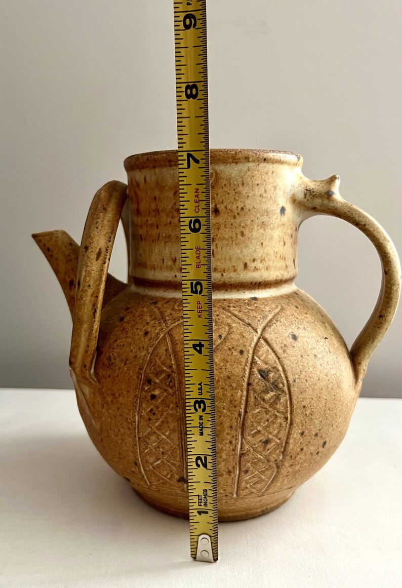May include: A brown ceramic teapot with a textured surface and a carved design. The teapot is 7 inches tall.