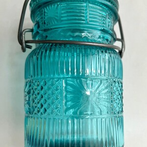 May include: A teal-colored glass jar with a decorative lid and a metal clasp. The jar features vertical and geometric embossed patterns. The lid has a textured rim. A stylish storage container for kitchen or home decor.
