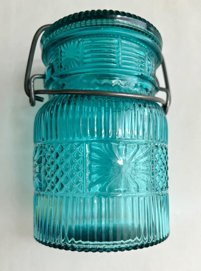 May include: A teal-colored glass jar with a decorative lid and a metal clasp. The jar features vertical and geometric embossed patterns. The lid has a textured rim. A stylish storage container for kitchen or home decor.