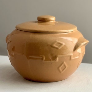 May include: A tan ceramic crock with a lid, featuring a diamond pattern and a textured surface. The crock has two small handles on the sides.