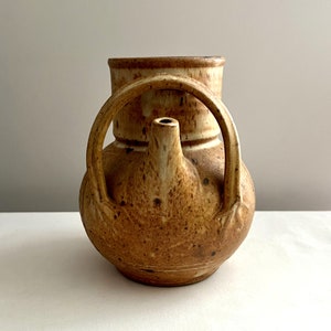 May include: A brown ceramic pitcher with a unique handle and a spout. The pitcher has a speckled glaze and a rustic look.