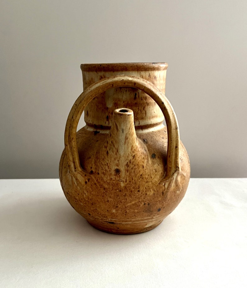 May include: A brown ceramic pitcher with a unique handle and a spout. The pitcher has a speckled glaze and a rustic look.