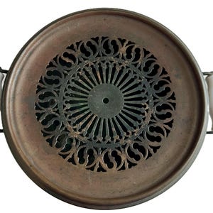 May include: Antique copper-toned metal food warmer with a decorative, openwork center. Features white ceramic handles on either side. The circular design includes a detailed, symmetrical pattern in the center.