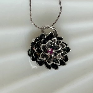 May include: A silver necklace with a pendant shaped like a dark flower with a small, round, red gemstone in the center. The flower pendant has detailed petals and is suspended from a delicate silver chain.