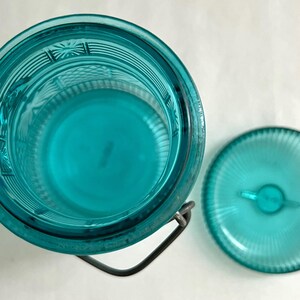 May include: An overhead view of a turquoise glass jar with a matching lid. The jar has a metal handle and a textured design. The lid is round with a small handle. The jar and lid are on a white surface.