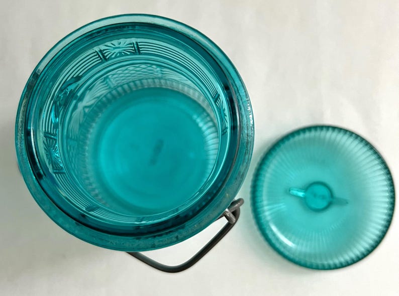 May include: An overhead view of a turquoise glass jar with a matching lid. The jar has a metal handle and a textured design. The lid is round with a small handle. The jar and lid are on a white surface.