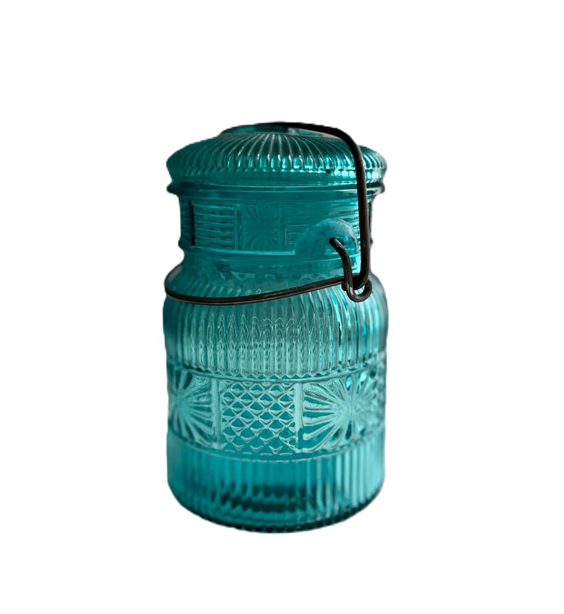 May include: A teal-colored glass jar with a lid and a metal clasp. The jar has a ribbed design on the lid and body, with a decorative pattern on the lower section. The jar is empty and sits against a white background.