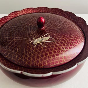 May include: A burgundy-colored decorative bowl with a matching lid. The lid features a gold geometric pattern and a detailed insect illustration. A small, red knob serves as a handle. The bowl has a scalloped edge.