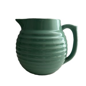 May include: A green ceramic pitcher with a ribbed design. The pitcher has a curved handle and a spout for pouring. The color is a muted, earthy green. The pitcher is likely used for serving beverages.