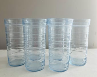 May include: Four light blue, ribbed drinking glasses. The glasses are cylindrical with horizontal ridges around the body. The glasses are empty and sit on a white surface against a gray background.