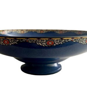 May include: A navy blue ceramic bowl with a pedestal base. The rim features a decorative border with red floral and white leaf accents. The bowl's interior is a lighter shade of blue, and the exterior is a darker, solid blue.