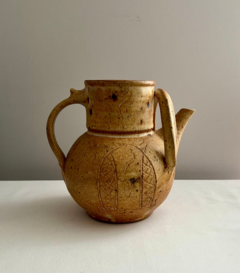 May include: A brown ceramic teapot with a unique design. The teapot has a rounded body with a spout and a handle. The handle is attached to the body with a small, decorative piece.