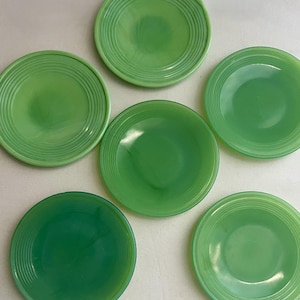 May include: Six jadeite green plates are arranged on a white surface. The plates are round with a slightly concave center and concentric ring detailing around the rim. The plates vary slightly in shade, from light to dark green.