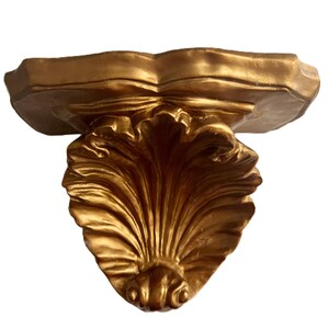 May include: A gold-colored wall shelf with a decorative, ornate design. The shelf features a scalloped top and a stylized, fan-shaped support. The shelf is made of a solid material and has a glossy finish.