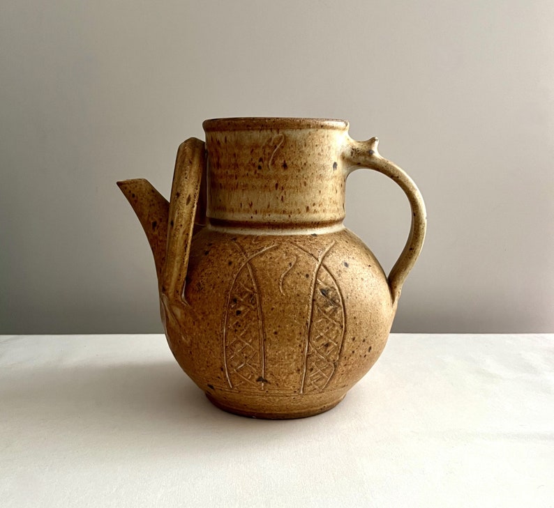 May include: A brown ceramic pitcher with a textured surface and a carved design. The pitcher has a handle and a spout.
