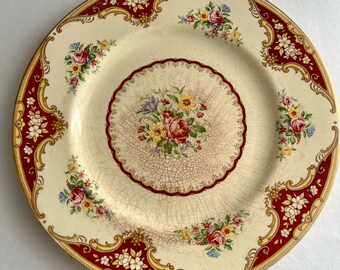 May include: A vintage floral dinner plate with a white background and a red and gold border. The plate features a delicate floral pattern with roses, leaves, and other flowers. The center of the plate has a smaller floral design with a cracked glaze effect.