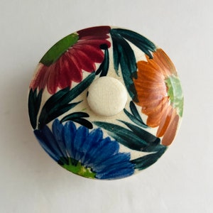 May include: A white ceramic lid with a floral design. The lid features three flowers in shades of red, orange, and blue, with green leaves. The center of the lid has a white knob.