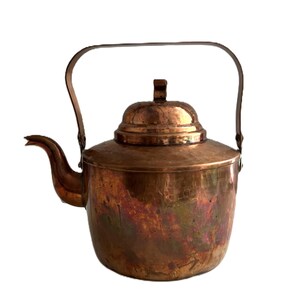 May include: A copper kettle with a lid and a curved handle. The kettle has a spout and a rounded body. The copper has a warm, reddish-brown color with some areas showing a slight iridescent sheen. The handle is attached to the sides of the kettle.