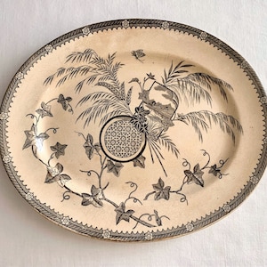 May include: An oval-shaped ceramic platter with a cream-colored background and a black and white floral design. The design features a central motif of a stylized flower with a geometric pattern, surrounded by ivy vines and a leafy branch.