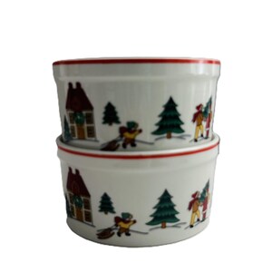 May include: Two white ceramic ramekins stacked on top of each other. Each ramekin has a red rim and is decorated with a winter scene featuring houses, evergreen trees, and people. The scene is in a cartoon style.