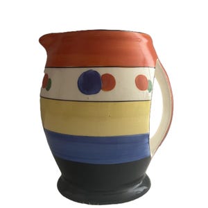 May include: A ceramic pitcher featuring horizontal bands of orange, white, yellow, blue, and black. The white band is decorated with a repeating pattern of blue and orange circular dots. The pitcher includes a curved handle and a pouring spout.