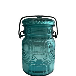May include: A teal-colored glass jar with a lid and a black metal handle. The jar has a ribbed design with decorative diamond and starburst patterns. The lid is also ribbed. The jar is likely for storage or decoration.