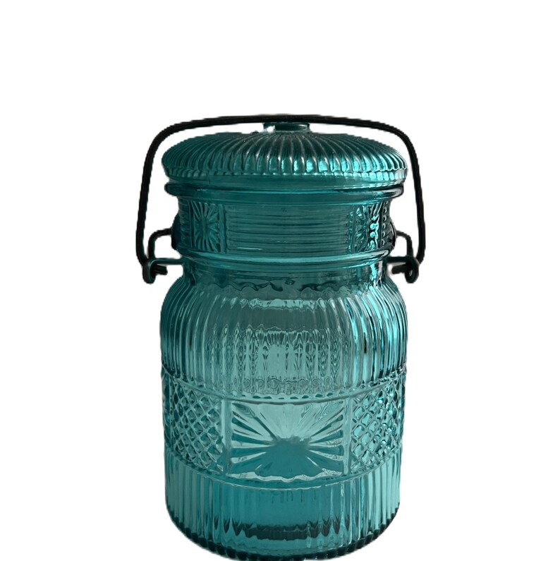 May include: A teal-colored glass jar with a lid and a black metal handle. The jar has a ribbed design with decorative diamond and starburst patterns. The lid is also ribbed. The jar is likely for storage or decoration.
