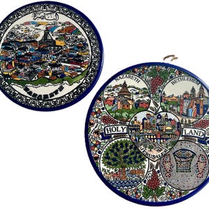 May include: Two decorative ceramic plates with blue rims. One plate features a detailed illustration of a city with text in a foreign language. The other plate has illustrations of Nazareth, Bethlehem, Jerusalem, and the Holy Land, with floral and grape designs.