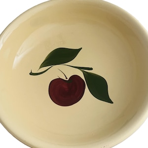 May include: A cream-colored ceramic bowl with a hand-painted design of a dark red cherry and two green leaves on a branch. The bowl has a slightly curved rim and a smooth surface.