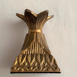 May include: A gold-colored, ornate candle holder with a detailed, leaf-like design. The base is a wide, triangular shape, and the top has a stylized, petal-like design. The candle holder is made of metal and has a matte finish.