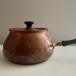 May include: A vintage copper pot with a wooden handle and a lid. The pot has a worn, aged look and is perfect for melting chocolate or cheese.