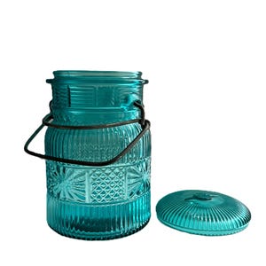 May include: A teal glass jar with a decorative pattern and a metal handle. The jar has a matching lid and is designed with vertical and starburst patterns. The jar is empty and sits on a white background.